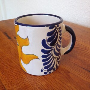 May include: A ceramic mug with a white base, featuring a yellow sun design and blue floral patterns. The rim and handle are dark blue. The mug is on a wooden surface.