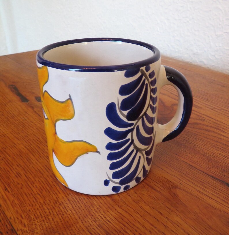 May include: A ceramic mug with a white base, featuring a yellow sun design and blue floral patterns. The rim and handle are dark blue. The mug is on a wooden surface.