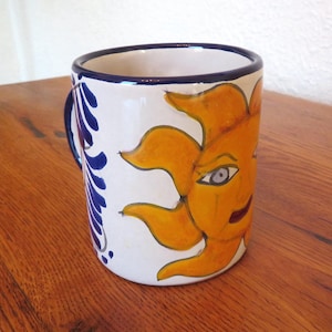 May include: White ceramic mug with a hand-painted design of a smiling sun with orange and yellow rays. The mug has a blue handle and a blue and white floral pattern on the side. The rim is trimmed with blue.