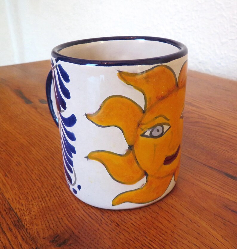 May include: White ceramic mug with a hand-painted design of a smiling sun with orange and yellow rays. The mug has a blue handle and a blue and white floral pattern on the side. The rim is trimmed with blue.