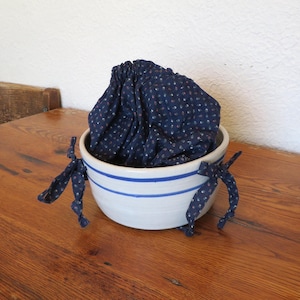 May include: A white ceramic bowl with two blue stripes and a navy blue fabric with a small red and white polka dot pattern. The fabric is gathered inside the bowl, and blue fabric ties are attached to the bowl.