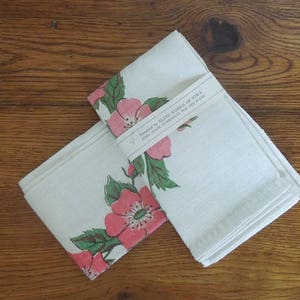 Pu&ograve; includere: Due asciugamani da t&egrave; in lino bianco con un motivo floreale rosa. Gli asciugamani sono piegati e impilati uno sopra l'altro. L'asciugamano superiore ha un'etichetta che recita "Handmade with Love in the USA".