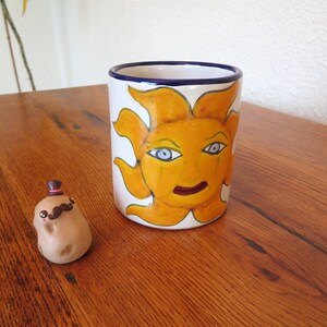 May include: A white ceramic mug with a hand-painted sun design in yellow, orange, and blue. A small potato figurine with a mustache and top hat is next to the mug. The mug is approximately 4 inches tall.