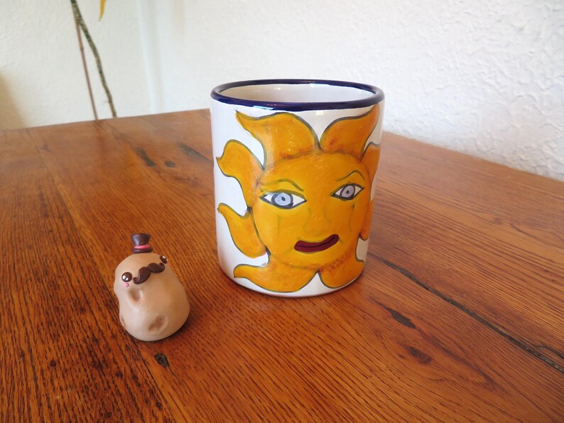 May include: A white ceramic mug with a hand-painted sun design in yellow, orange, and blue. A small potato figurine with a mustache and top hat is next to the mug. The mug is approximately 4 inches tall.