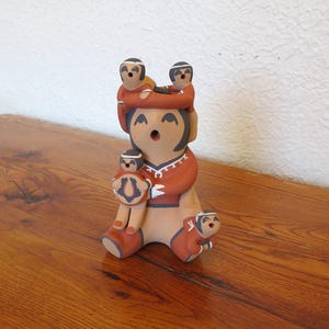 May include: A handcrafted ceramic figurine depicting a mother figure with four children. The figurine is painted in shades of tan, brown, and red, with black accents. The children are positioned on the mother's head, arms, and lap. The figurine is a decorative item.
