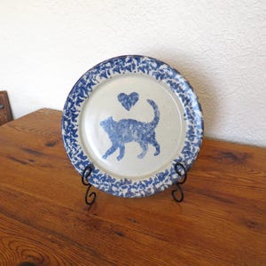 May include: A ceramic plate with a blue cat and heart design. The plate has a white center with a blue cat silhouette and heart. The rim features a blue floral pattern. The plate is displayed on a black metal stand.