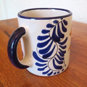 May include: White ceramic mug with a dark blue handle and rim. The mug features a blue floral design with yellow accents. The mug is a standard size for coffee or tea.