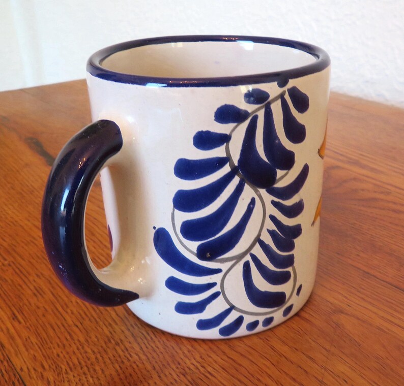 May include: White ceramic mug with a dark blue handle and rim. The mug features a blue floral design with yellow accents. The mug is a standard size for coffee or tea.