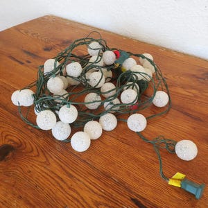 May include: A string of white globe lights with green wiring and a green plug. The lights are clustered together on a wooden surface. The globe lights are round and have a textured surface.