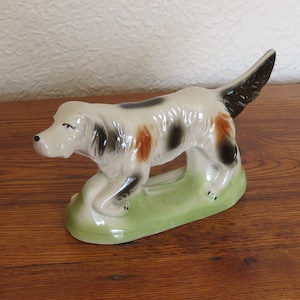 Puede incluir: Figura de cerámica de un perro en posición de caminar. El perro tiene pelaje blanco con manchas negras y marrones, una cola negra, y está sobre una base verde. La figura está sobre una superficie de madera.