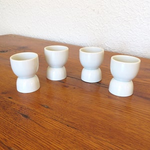May include: Four white ceramic egg cups on a wooden table. The egg cups have a two-tiered design.
