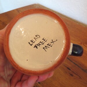 May include: A ceramic mug with a white interior and a brown rim. The handle is dark blue with a white stripe. The words "LEAD FREE MEX." are written in black on the bottom of the mug.