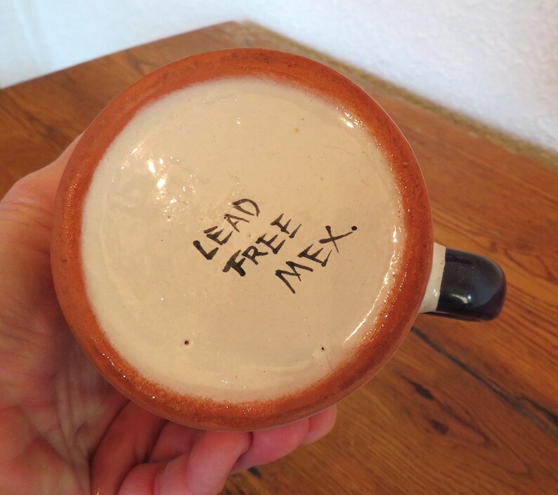 May include: A ceramic mug with a white interior and a brown rim. The handle is dark blue with a white stripe. The words "LEAD FREE MEX." are written in black on the bottom of the mug.