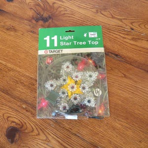 May include: A Christmas tree star topper with 11 lights, packaged in a clear plastic case. The star has a yellow center and silver tinsel-like edges. The packaging is green and white and includes the text "11 Light Star Tree Top" and the Target logo.