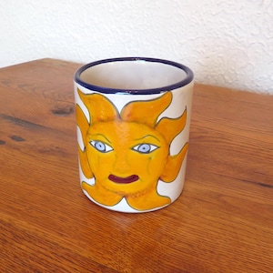 May include: A cylindrical ceramic mug with a hand-painted sun face design. The sun has yellow rays, blue eyes, and a red mouth. The rim is trimmed with a blue band. The mug is on a wooden surface.
