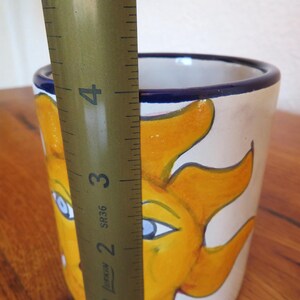 May include: A hand-painted ceramic mug featuring a smiling sun design in yellow and orange against a cream background. The mug has a blue rim and measures approximately 4 inches in height.