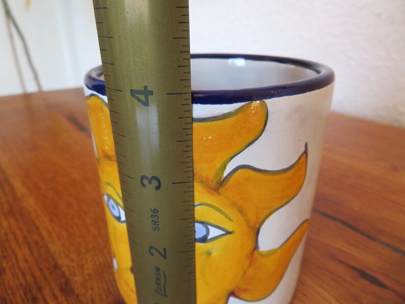 May include: A hand-painted ceramic mug featuring a smiling sun design in yellow and orange against a cream background. The mug has a blue rim and measures approximately 4 inches in height.
