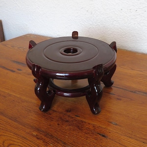 May include: A dark reddish-brown wooden display stand with a round top and ornate legs. The stand has a central hole and is designed to hold a decorative object. The stand is approximately 6 inches in diameter.
