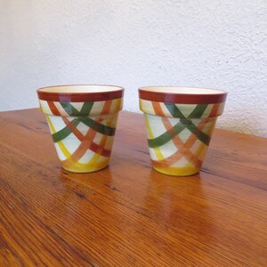 May include: Two ceramic flower pots featuring a hand-painted crisscross pattern in shades of orange, yellow, and green. Each pot has a solid dark orange rim and a cream-colored interior, with a tapered shape that widens toward the top opening.