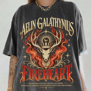 May include: A charcoal grey short-sleeve t-shirt featuring a graphic of a deer wearing a crown surrounded by flames and stars. The text reads AELIN GALATHYNIUS FIREHEART, followed by the quote: SHE WAS FIRE, AND LIGHT, AND ASH, AND EMBERS.