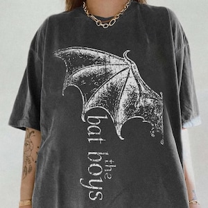 Op de afbeelding: Een antracietgrijs t-shirt met korte mouwen en een verweerde witte afbeelding van een vleermuisvleugel en de tekst "bat the boys" verticaal gedrukt. Het shirt heeft een ronde hals en een relaxte pasvorm met een vintage gewassen textuur.