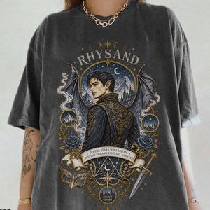 May include: A charcoal grey short-sleeve t-shirt featuring a graphic print of a winged figure with pointed ears. The design includes the text "RHYSAND" and "TO THE STARS WHO LISTEN AND THE DREAMS THAT ARE ANSWERED," along with a dagger, mask, and roses.