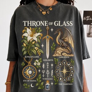 以下が含まれることがあります： Throne of GlassのテキストがプリントされたチャコールグレーのTシャツ。デザインには、白いユリ、Goldrynという剣、Abraxosというドラゴン、Eye of Elenaというペンダント、Wyrdmarks、円形エンブレムが描かれています。