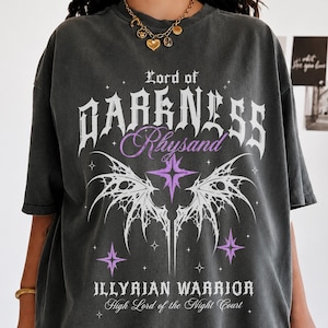 Op de afbeelding: Een antracietgrijs t-shirt met korte mouwen en witte en paarse grafische tekst met de tekst Lord of Darkness Rhysand Illyrian Warrior High Lord of the Night Court, vergezeld van een witte vleugelillustratie en paarse steraccenten.