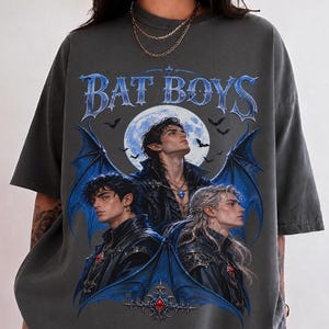 Peut inclure: Un t-shirt gris anthracite à manches courtes avec une impression graphique portant le texte "BAT BOYS" en lettres bleues. Le motif représente trois personnages aux cheveux sombres, des ailes de chauve-souris bleues et une pleine lune.