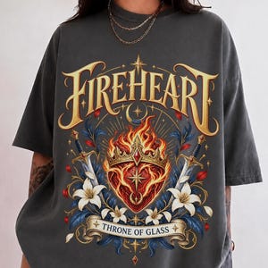 May include: A charcoal grey short-sleeve t-shirt featuring a graphic print with the text FIREHEART and THRONE OF GLASS. The design includes a flaming heart wearing a crown, flanked by two swords and white lilies, all rendered in gold and red tones.