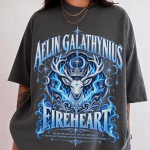 May include: A charcoal grey short-sleeve t-shirt featuring a graphic of a white stag wearing a crown, surrounded by blue flames, stars, and crescent moons. The text reads AELIN GALATHYNIUS FIREHEART and includes a quote at the bottom.