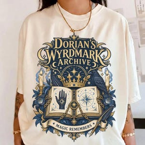 Puede incluir: Una camiseta color marfil con un estampado gráfico que incluye el texto "Dorian's Wyrdmark Archive" y "Magic Remembers". El diseño muestra un libro abierto, una corona, un cuervo, velas, flores y un símbolo de mano en tonos azules y dorados.