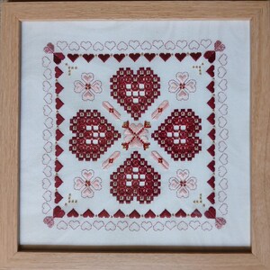 May include: A framed cross-stitch embroidery featuring a design of red hearts with a white background. The hearts are arranged in a symmetrical pattern with a central heart surrounded by four smaller hearts.