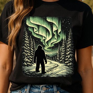 May include: Black t-shirt featuring a graphic of a Bigfoot silhouette standing on a snowy path, beneath a vibrant green aurora borealis. The design includes detailed pine trees and a starry night sky.