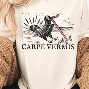 May include: Cream-colored t-shirt featuring a black and white graphic of a bird with a worm in its beak, set against a landscape with a rising sun. The text "CARPE VERMIS" is printed below the graphic.
