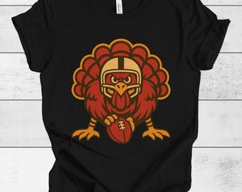 Puede incluir: Camiseta negra con un pavo de dibujos animados que lleva un casco de fútbol americano y sostiene un balón de fútbol. El pavo es de color rojo, marrón y dorado. La camiseta es de un material suave.