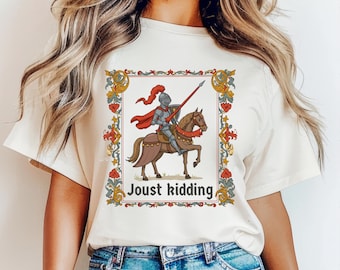 Pode incluir: Uma camiseta branca com uma ilustração de estilo medieval de um cavaleiro a cavalo. A ilustração é cercada por uma borda decorativa e o texto "Joust kidding" é impresso abaixo.