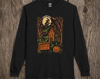 May include: A black long-sleeve t-shirt featuring a graphic print of a witch flying on a broomstick in front of a full moon, a house, trees, a cat, a pumpkin, and a sign that reads SALEM 400 and SALEM, MA EST. 1626 in orange and yellow tones.