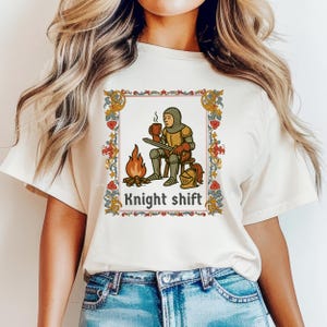 May include: Cream-colored t-shirt featuring a medieval-style illustration of a knight sitting by a fire, holding a mug. The image is framed with a decorative border and the text "Knight shift" is below the illustration.