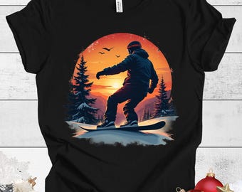 Könnte beinhalten: Ein schwarzes T-Shirt mit einem Grafikdruck eines Snowboarders, der vor einem Sonnenuntergang silhouettiert ist. Der Snowboarder trägt einen Helm und fährt einen Berg hinunter. Die Grafik ist von Bäumen und Schnee umgeben.