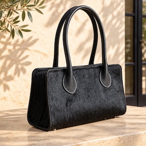 Könnte beinhalten: Eine rechteckige schwarze Handtasche mit einer texturierten Oberfläche aus Fell. Die Tasche verfügt über zwei abgerundete Tragegriffe, die mit kreisförmigen Lederaufnähern befestigt sind, und einen strukturierten Boden mit sichtbaren Metallfüßen.
