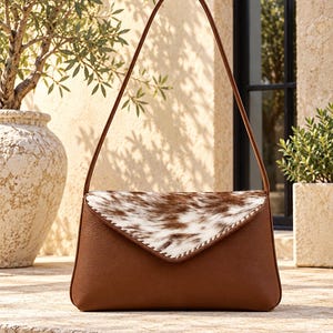May include: A brown leather shoulder bag featuring a triangular flap with a white and brown cowhide pattern. The flap is accented with decorative whipstitching along the edges, and the bag includes a single thin shoulder strap.