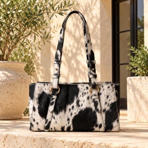 May include: A rectangular handbag featuring a black and white cowhide pattern with a textured, hairy finish. The bag includes two vertical straps attached to gold-toned metal rings, leading to two shoulder straps with gold-toned hardware accents.