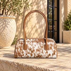 May include: A cylindrical handbag featuring a brown and white cowhide pattern with smooth tan leather trim and two matching tan leather shoulder straps. The bag has a top zipper closure and a structured rectangular base with rounded edges.