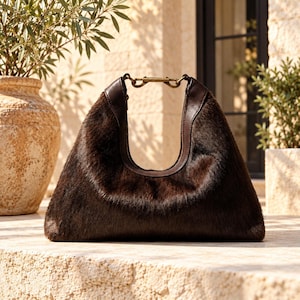 May include: A dark brown hobo-style handbag crafted from textured faux fur material. The bag features smooth dark brown leather trim along the top opening and a metallic gold-toned hardware clasp connecting the two sides of the handle.