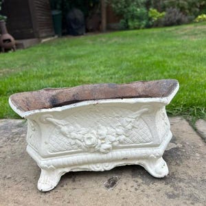 May include: A rectangular off-white planter with a weathered, rusted interior. The exterior features a raised floral relief pattern, a lattice-style texture, and four ornate feet. The planter has a wide, open top and a sturdy, heavy-duty construction.