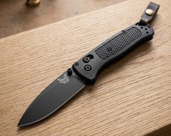 May include: A folding pocket knife with a matte black blade and a textured black handle. The blade features a small butterfly logo near the base. A dark brown leather lanyard with a brass-colored metal snap button is attached to the end.