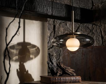 May include: A pendant light fixture featuring a central spherical white glass bulb encased within a larger, flattened, translucent dark gray glass shade. The fixture is suspended by a thin metallic rod and emits a soft, warm glow.
