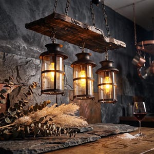 May include: A rustic chandelier featuring three cylindrical glass lanterns with dark metal frames suspended from a horizontal piece of weathered, dark brown wood by metal chains, each lantern containing a glowing light bulb.