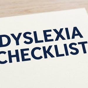 Op de afbeelding: Een witte rechthoekige kaart met de donkerblauwe tekst DYSLEXIA CHECKLIST in een vet schreefloos lettertype. Een blauwe en doorzichtige plastic balpen ligt rechts van de kaart op een lichtgekleurd houten oppervlak.